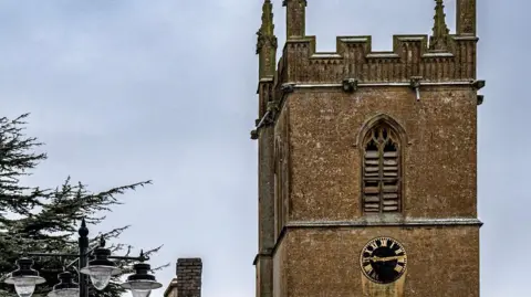 Mike Schoen An old church with a black and gold clock on the side. The sky is grey and cloudy. There is the side of a tree in the far left and the top of a lamp post. 