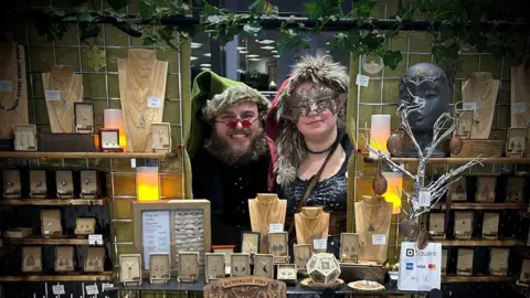 Handout Dave and Sandra Hubble stand behind the Wyrdwood jewellery stall. They are wearing elf hats for Christmas and the stall is decorated with evergreens. Jewellery stands and boxes are laid out showing the items they have made.