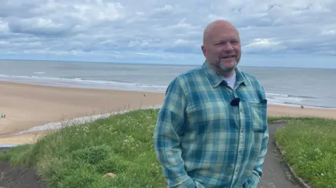 Jason Camis is standing with the large sweep of Longsands beach behind him. The sea is mostly calm, apart from wavelets near the shore, and the sky is blue but filled with white clouds. He is wearing a blue checked shirt, and has a receding hairline and stubble. 