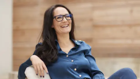 Jocelyn Penque Wearing a blue-satin blouse and with dark hair Jocelyn Penque smiles, while seated and looking to the side of the shot.
