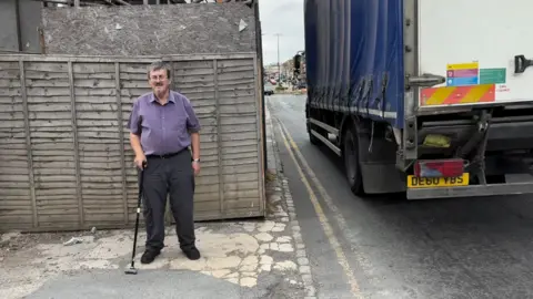 Martin Heath/BBC Giles du Boulay standing with a walking stick on a pavement beside a construction site enclosed by wooden fencing and a black site cabin. Scaffolding rises behind the fence, while warning signs, an assembly point sign and a construction traffic sign are attached to the barriers. A road with cars and office buildings runs alongside the site under an overcast sky.