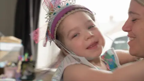 A little girl wearing a toy crown, with her arms around her mum's neck who is slightly out of frame.