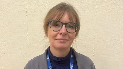 Melanie Pittock Melanie Pittock is looking at the camera against a cream wall. She has light brown hair which is tied back and is wearing dark-rimmed spectacles. She has a dark blue jumper on and a greay jacket. 