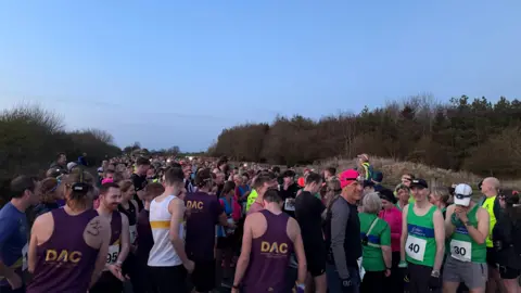 Ollie Rawlinson/BBC A large group of people standing in the nature reserve which is surrounded by leafless trees on either side. Most of them are wearing running vests and there are a range of colours from purple to green. It looks like it is dusk.