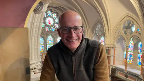 A bald man in glasses and a brown fleece sits in a high alcove in a church.