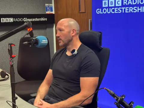 A man talking into a microphone with a purple screen in the background and BBC Radio Gloucestershire branding. 