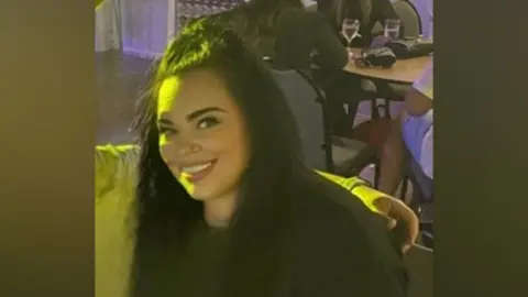 Laura Whybrow A woman smiles at the camera. She has long dark hair, some of which has been tied back. A person, who is off camera, rests their arm around her shoulders. People at a table can be seen behind her. 