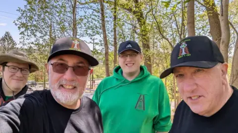 Duffy's Cut Project Frank Watson has a grey beard and is wearing a dark long-sleeved top, sunglasses and a Duffy's Cut baseball cap. Bill Watson is clean-shaven and is also wearing a dark top and Duffy's Cut baseball cap. Two other men are also in the picture. Both are wearing glasses, one is wearing a baseball cap and the other a hat.