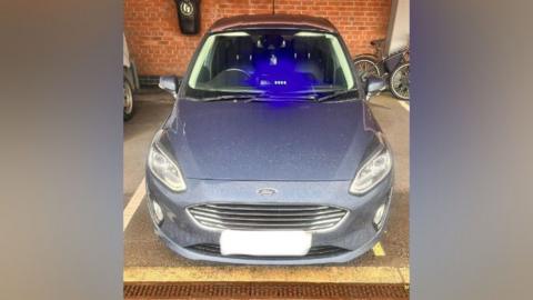 A picture of the car issued by the police. It shows a silver/grey Ford parked in what appears to be a covered parking space. The blue light is clearly visible flashing in the front seat. 