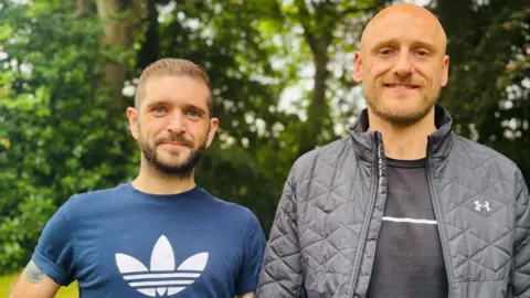 Kieran Hutcheon and Dan Bickmore, two men in their 30s smiling at the camera, in front of trees.