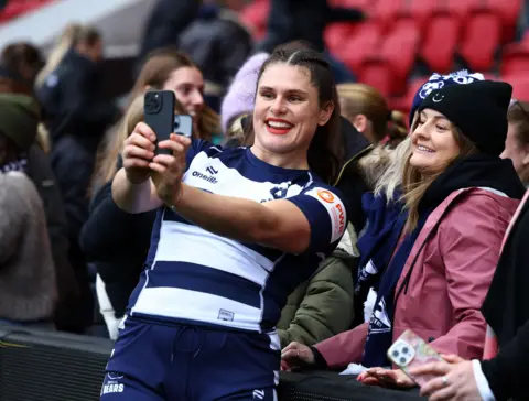 Ilona Maher poses for selfies with fans in a sports stadium. 