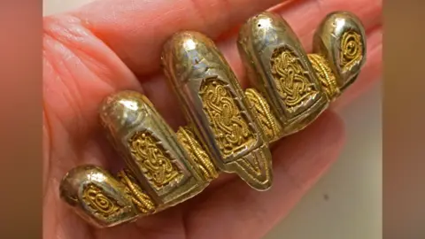 The Portable Antiquities Scheme A close-up of a Viking-era silver and gold sword pommel resting on a hand. It is made up of five lobes, which decrease in width from the tallest one in the middle. Each of the lobes has an inset panel with an interlocking pattern of gold wire on a gold panel beneath. More gold wire is woven between the lobes. 