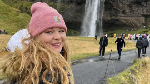 A young woman wearing a pink woollen hat, looks over her shoulder and smiles into camera in front of a waterfall