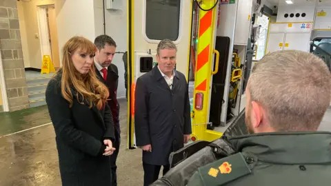Simon Dedman/BBC Sir Keir Starmer and Angela Rayner talking to a man at the rear of an ambulance that has its doors open. They are outside the Princess Alexandra Hospital