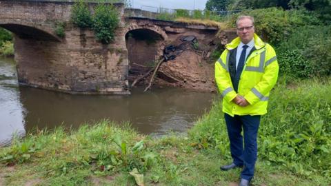  Councillor Marc Bayliss in a hi vis jacket standing in front of a collapsed bridge