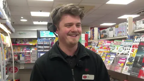 BBC/Aaron Outram A young man with brown hair in a post office uniform, stood in the magazine aisle