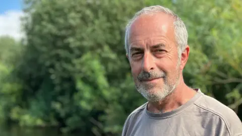 A man wearing a grey shirt looks towards the camera, with a stretch of river visible in the background.