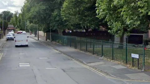 A white van drives on Clifford Street in Wolverhampton. Double yellow lines can be seen on the side of the road, alongside the street sign. Green railings and grass can be seen to the right of the image. Other cars can be seen in the background.