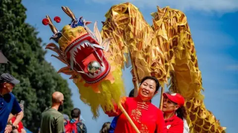 Faustina Yang Faustina, a woman wearing a red traditional Chinese outfit, holding a yellow and orange paper dragon.