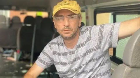 A man wearing a yellow cap and a grey t-shirt sitting in the back of a van.