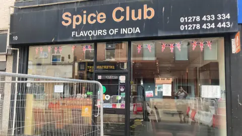 The picture shows the outside of The Spice Club in Bridgwater.  The sign is black with Spice Club written in orange paint.  The sign also says Flavours of India. 