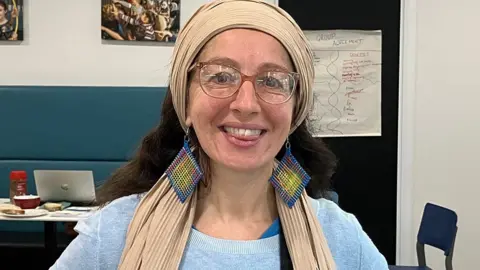 Head and shoulders of Jen Rankin standing smiling in a dining room. She has long dark wavy hair and glasses and is wearing a beige scarf round her hair and a blue jumper and long square earrings in a woven blue , yellow and red pattern
