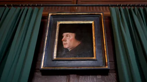 Gareth Fuller/PA A framed portrait of Thomas Cromwell on a dark wooden wall. The frame is dark brown wood with gold trim. To the left and right of it are green curtains. A black curtain rail runs above the portrait