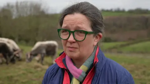BBC/Judy Hobson Julia Aglionby has brown hair and green framed glasses. She is wearing a blue coat with a red and a blue top inside. She has a colourful scarf. She is standing in a field with trees and long-horned cattle behind her.