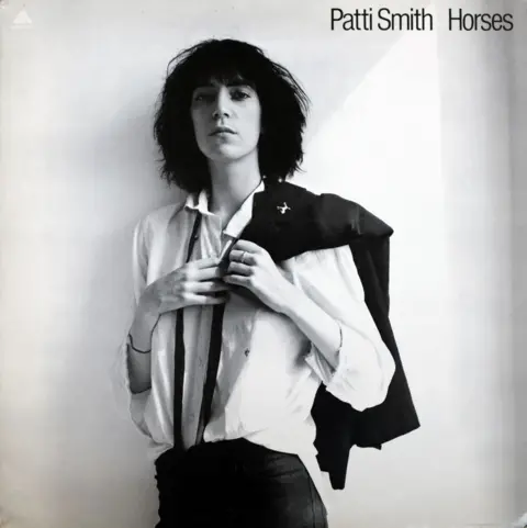 Patti Smith/Robert Mapplethorpe Foundation Patti Smith in white shirt with a dark ribbon hanging around her collar, and  holding a dark jacket over her shoulder