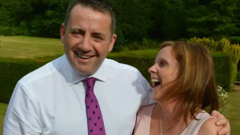 Supplied John Hunt is in a white shirt and wearing a purple tie with black spots. His wife Carol is facing him and laughing. They're standing outside on grass.