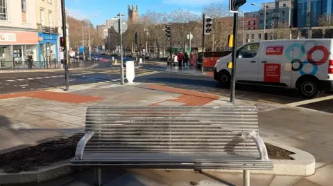 The bench seen from the front. Behind it is a view of Bristol city centre, with shops and a church.