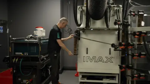 A man stands at a huge film projection machine that says IMAX on the side 