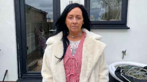 Suzie Cavadino has long, black hair and wears a white coat on top of a red-and-white gingham top. She is standing in front of her home and looks sad.