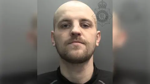 A custody photograph of a man with a shaved head and a brown beard and moustache. He is wearing a black top.