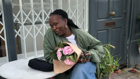 BBC Young girl sits at a small table in front of a cafe holding pink flowers and smiling.