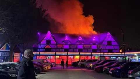 Gabby Cantareli The outside of Pink Punters, a night club in Fenny Stratford. There are purple neon lights and smoke and orange flames coming from the top of the building.