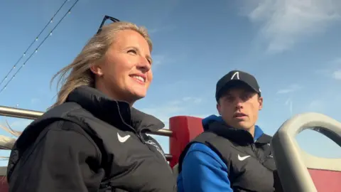 BBC A man and a woman sat on the top deck of an open-top bus. The man is wearing a black cap and looking directly at the camera. The woman is looking into the distance. 