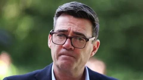 Prime Minister Andy Burnham wears dark-rimmed glasses and a blue suit.