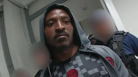 A still from a bodyworn police camera showing Temitope Adeyinka wearing a sports jersey and a grey hoodie, with the hood over his head. Police officers are standing around him, in a corridor, with their faces blurred out.