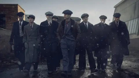 Peaky Blinders: The Immortal Man - a group of nine men in 1940s attire walking in an industrial area