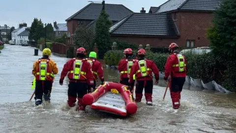 Shropshire Fire and Rescue Service A group of men in red jackets and yellow helmets pulling a red boat through a flooded street with a number of houses in the background