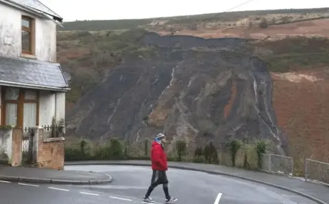 A man in red top and black leggings walks across a road with the damage from an old tip sliding down the mountain in the background