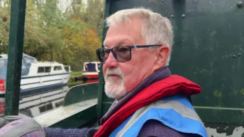 BBC/Nathan Turvey A picture of Bill Bartholomew on board one of the charity's boats, he has white hair and a white beard and is wearing a red life jacket.
