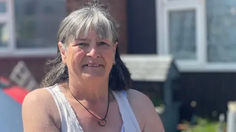 A woman in a white top is standing in her front garden with her home in the background. She has grey hair, tied back and wears a black necklace with a ring on it.