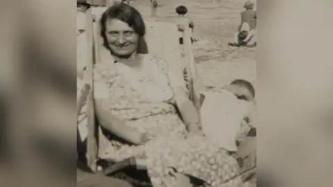 Avon and Somerset Old photograph of Louisa Dunne. She has dark hair and is sat in a deck chair wearing a floral dress. She is looking at the camera and smiling.