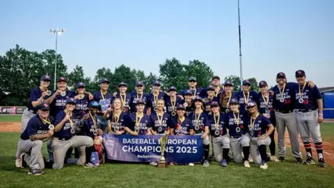 A group of women stand for a team photo in their Great Britain navy baseball kits and baseball hats. They are all wearing gold medals and have the trophy in front of them and a banner that says Baseball European Champions 2025.