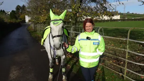 Amanda Wallace stands next to Murphy, a grey horse wearing a hi-vis sheet in the colour of Cumbria Police. Wallace is looking into the camera. She has brown hair and is wearing a hi-vis police jacket. They are standing in front of a field.