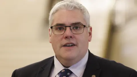Liam McBurney/PA Wire  Gavin Robinson wears glasses. He has a black suit on with a purple shirt and a tie. The background is blurred. 