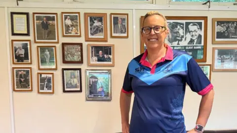 PA Media A woman wearing a cricket top is smiling with framed photos behind her