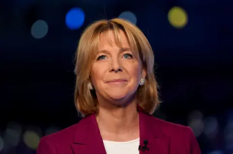 PA Media Hazel Irvine smiling gently and looking up with a blurred tv studio background. She is wearing a white t-shirt, sparkly earrings and a deep pink tailored jacket.